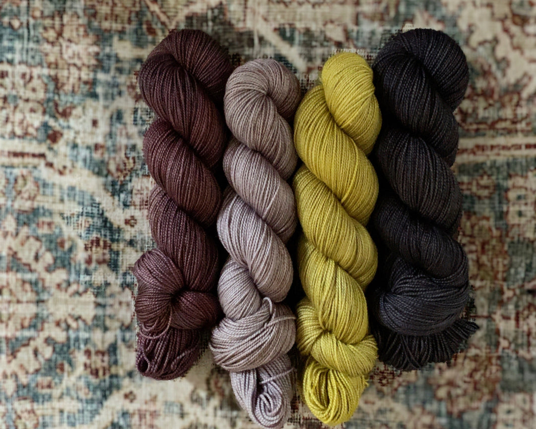 Four skeins of vixen yarn in Come on a My House, Minnie Pearl, Lemon Squeezy, and Raven's Wing on a patterned rug.